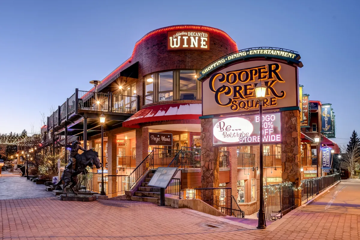 A lively exterior shot of Cooper Creek Square, featuring its welcoming sign, outdoor seating, wine shop, festive decorative lighting, and a crowd enjoying the vibrant atmosphere. The scene is animated with people engaged in shopping, dining, and entertainment—reflecting the energetic heart of downtown Winter Park.