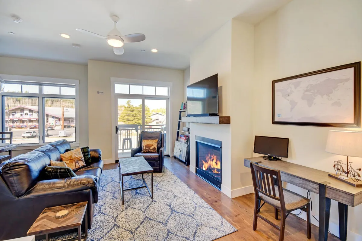 A cozy living area with a plush leather sofa, a fireplace, a desk setup for remote work, and a large window offering scenic mountain views. The atmosphere is inviting and versatile, suitable for relaxation, socializing, or enjoying the surroundings.