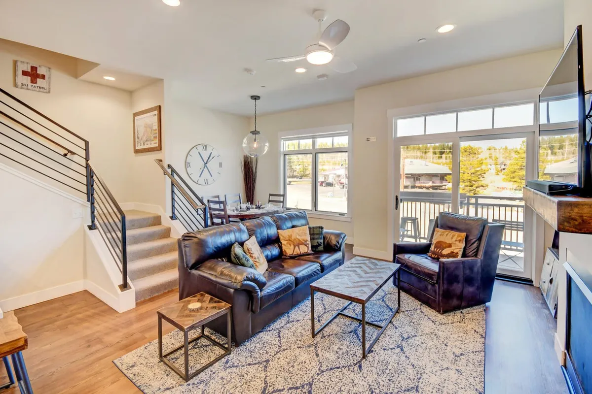 A warm, inviting living and dining area featuring a leather sofa, coffee table, large windows that let in natural light, and a glimpse of the adjacent dining space. Decorative wall art adds a cozy, personal touch, and the space feels ideal for game nights or relaxed evenings by the fire.