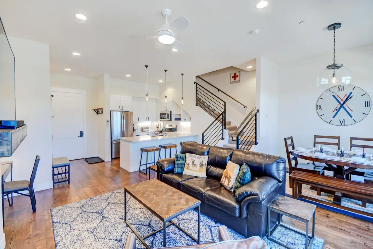 An open-plan view of the living room, dining area, and kitchen featuring a leather sofa, elegant dining table, kitchen island, and a staircase in the background. The design is contemporary and promotes effortless movement between social, cooking, and dining spaces.