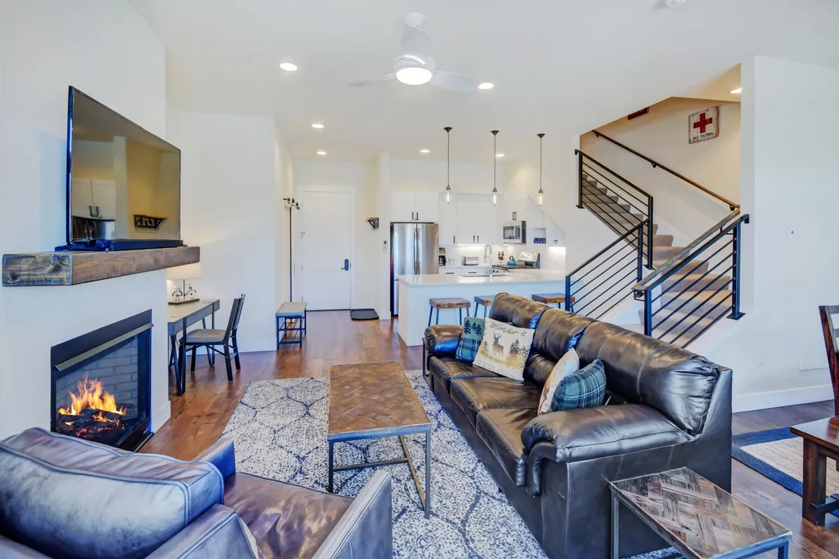The image showcases a warm, open-concept living space with a crackling fireplace, inviting sofa, modern kitchen area, and an elegant staircase. The mood is cozy and perfect for gathering, with spaces ideal for relaxing after mountain adventures, sharing meals, and enjoying quality time together.