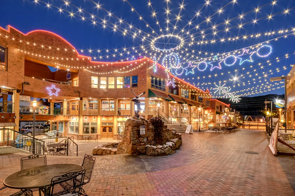 A festive, pedestrian-friendly street scene alive with the glow of decorative lights, a central stone fountain, and bustling storefronts. Outdoor seating and people socializing evoke the lively community and holiday spirit of Winter Park’s downtown area.