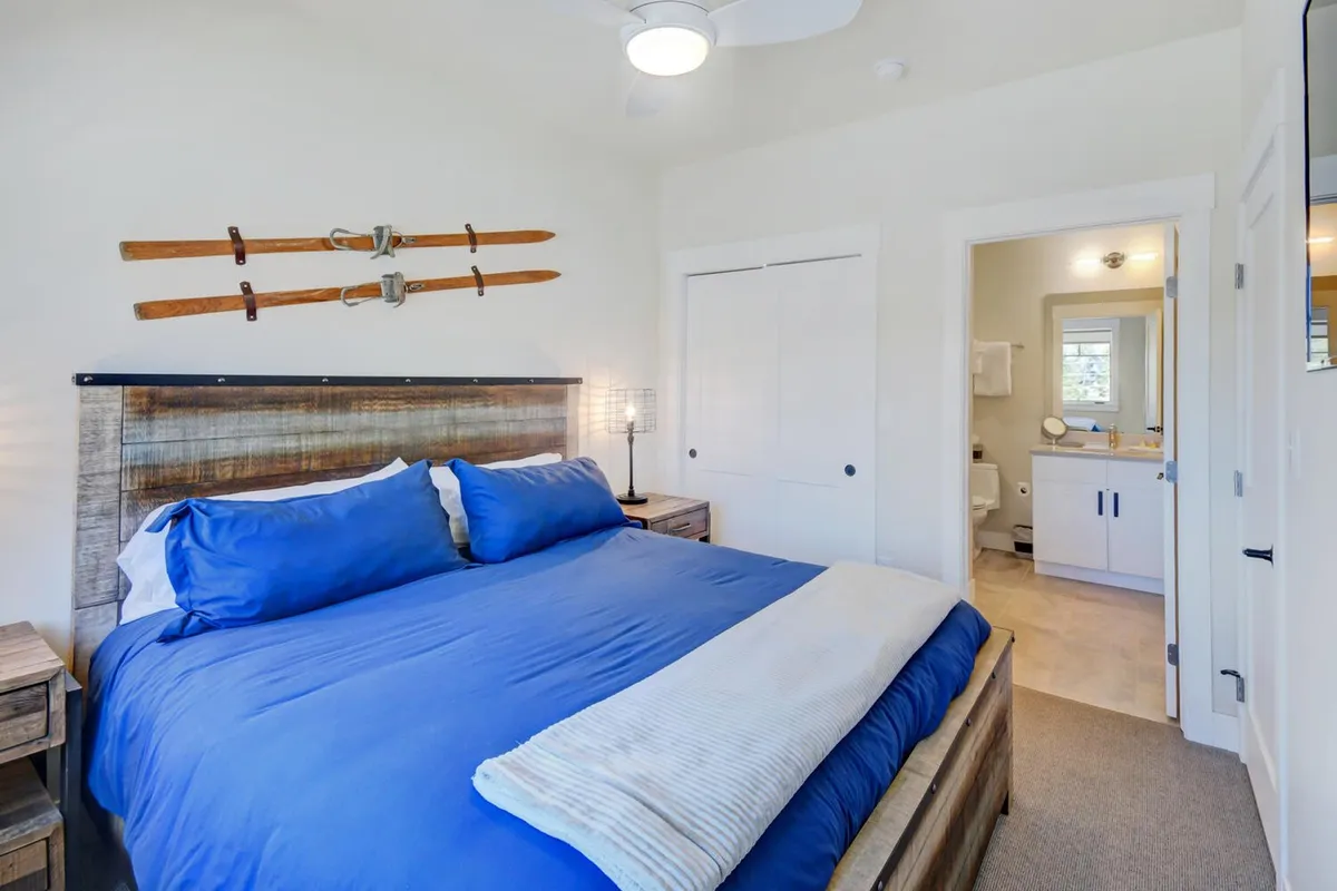 A spacious bedroom featuring a large blue king bed adorned with decorative pillows, wooden skis mounted on the wall as a unique accent, and a modern light fixture overhead. The setting is serene and invites relaxation, offering ample room for families or couples to unwind.