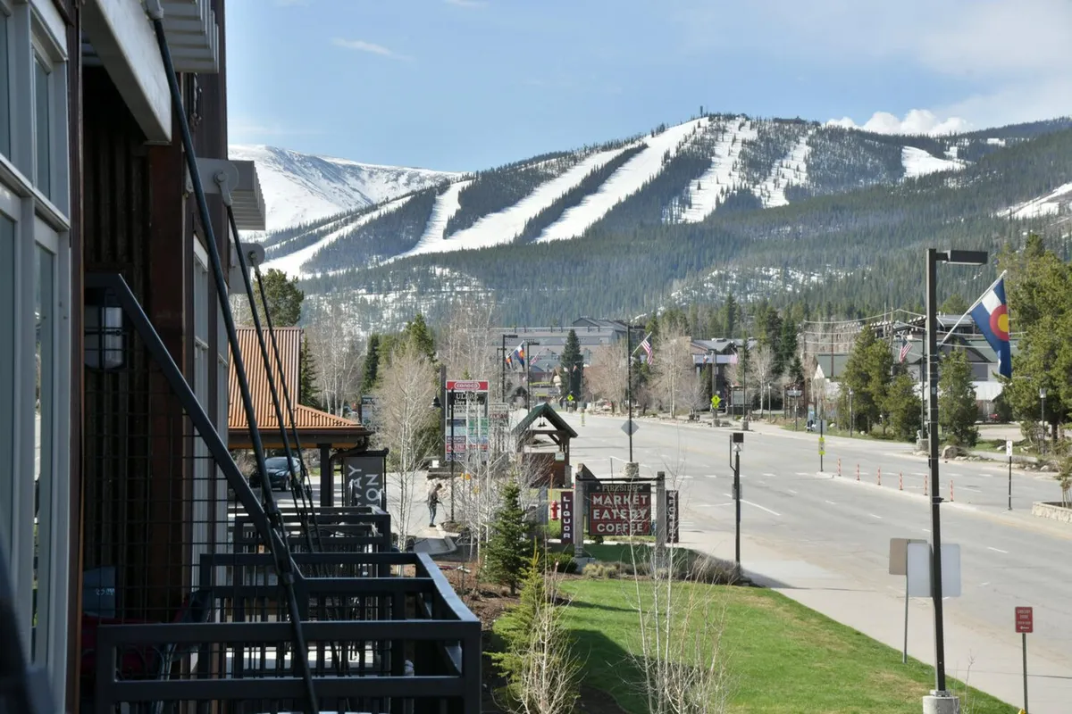 A lively Winter Park main street scene lined with charming storefronts, under the backdrop of snow-capped mountains. Balcony railings and street signs contribute to the feeling of being at the heart of the action, while the visible mountains hint at year-round adventure. The atmosphere is both adventurous and relaxed, with outdoor spaces suitable for gathering or setting off on an excursion.
